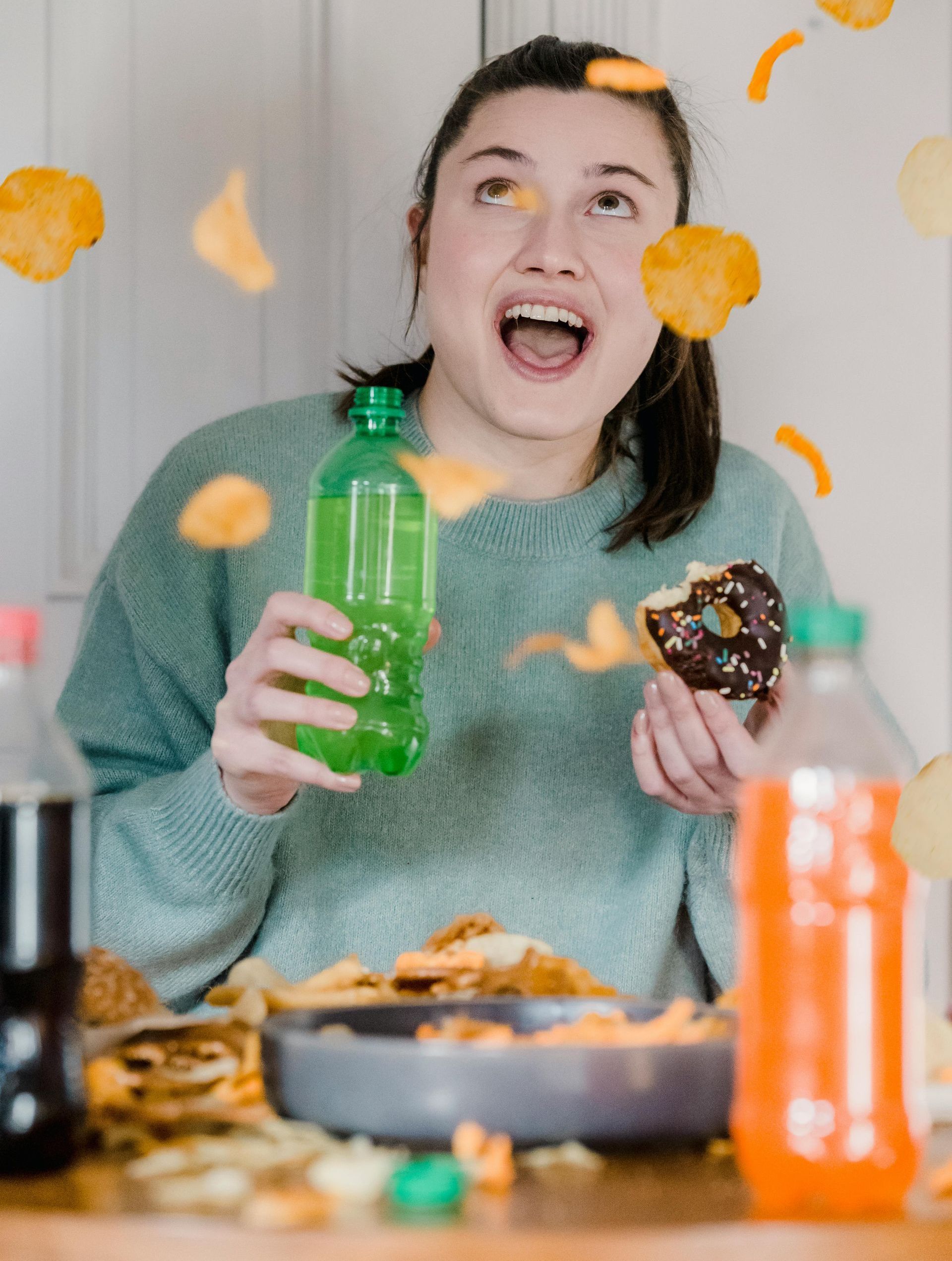 Woman with open mouth, holding soda, donut, chips falling. Snacks on table.