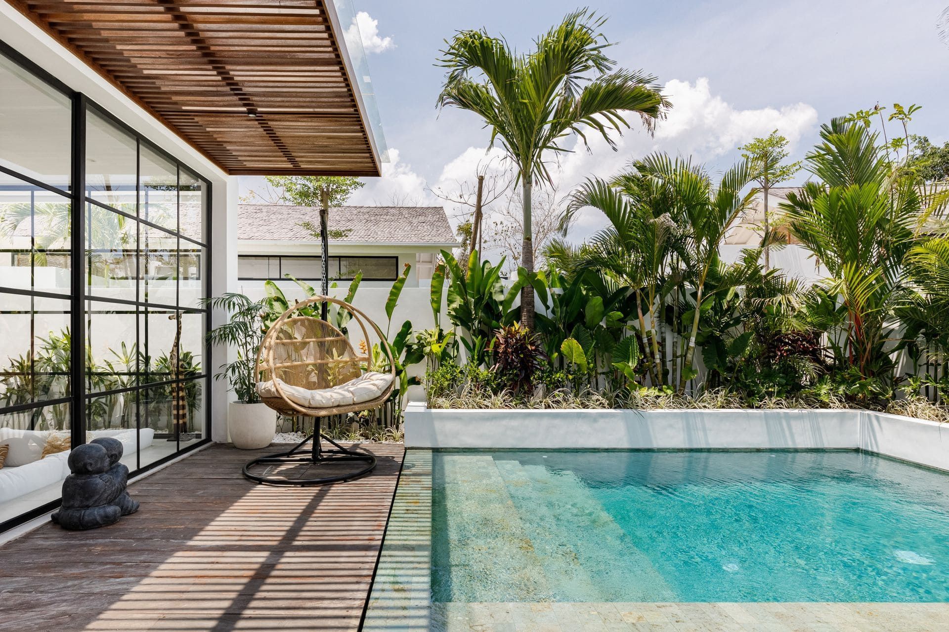 Modern tropical patio with a pool, wicker chair, and lush greenery beside a glass-walled house