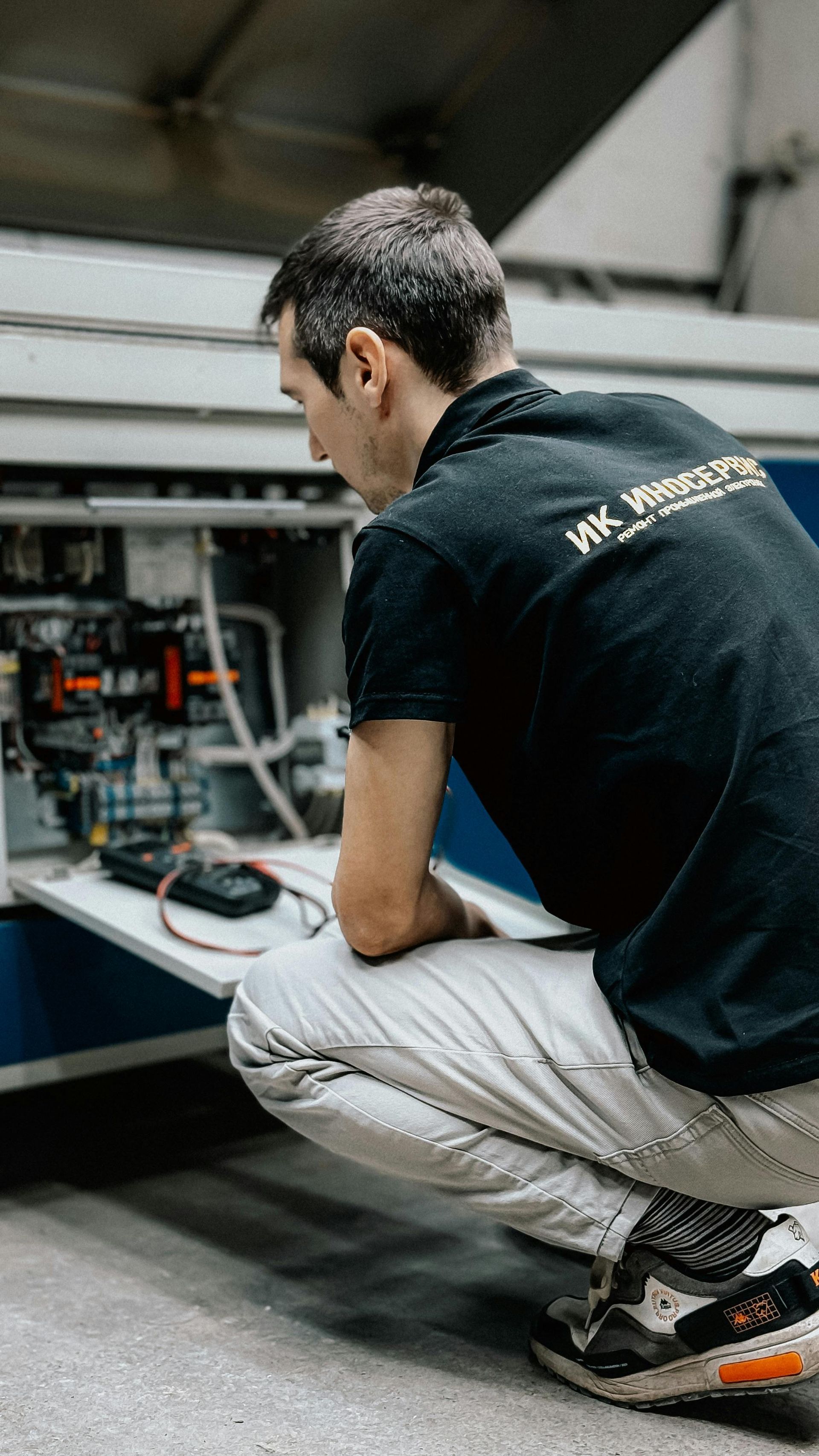 Hombre inspeccionando maquinaria, agachado frente a un panel de control abierto. Camisa negra y pantalones color canela.
