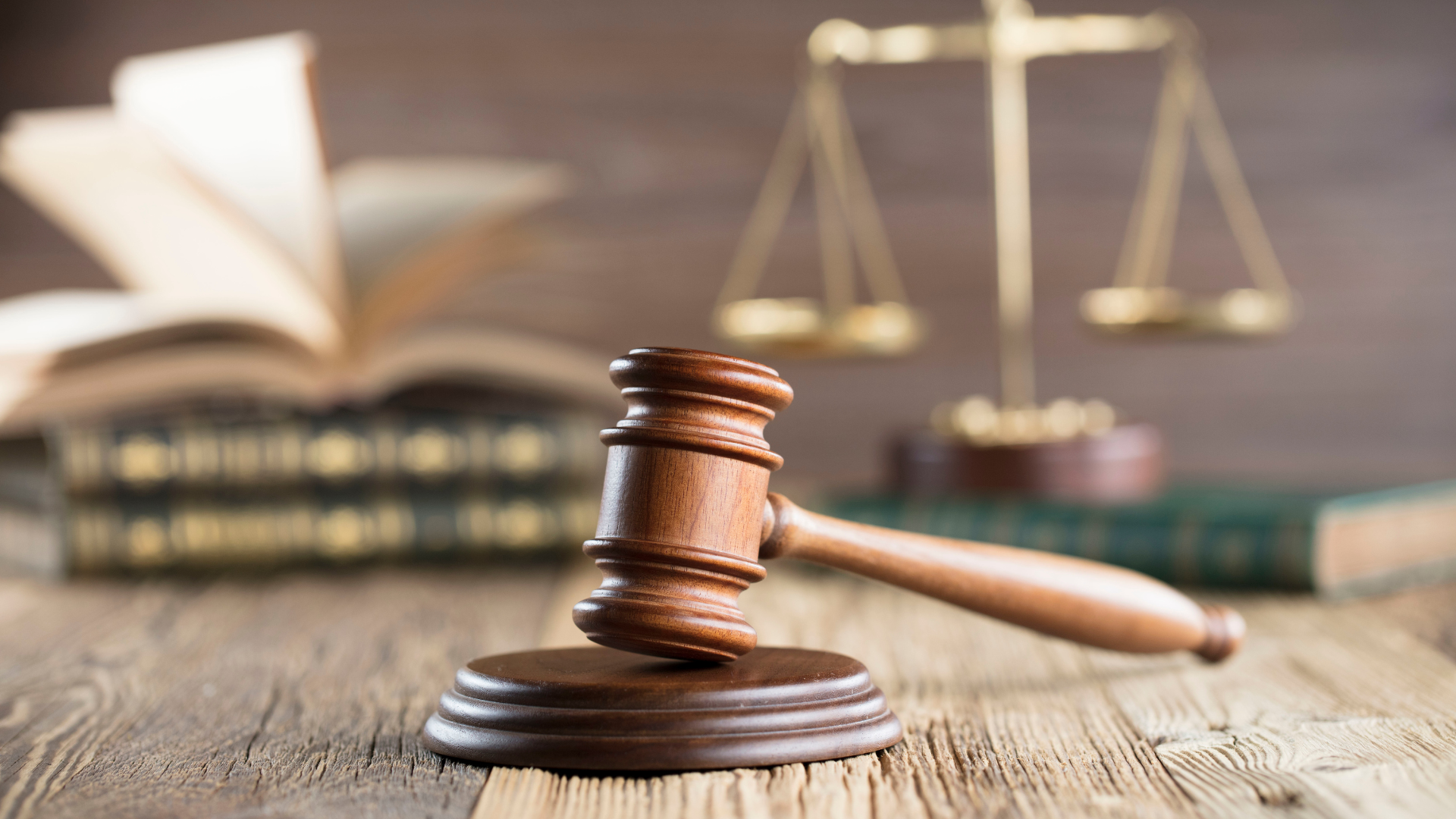 Wooden gavel on a wooden base, scales of justice, and law books on a wooden surface.