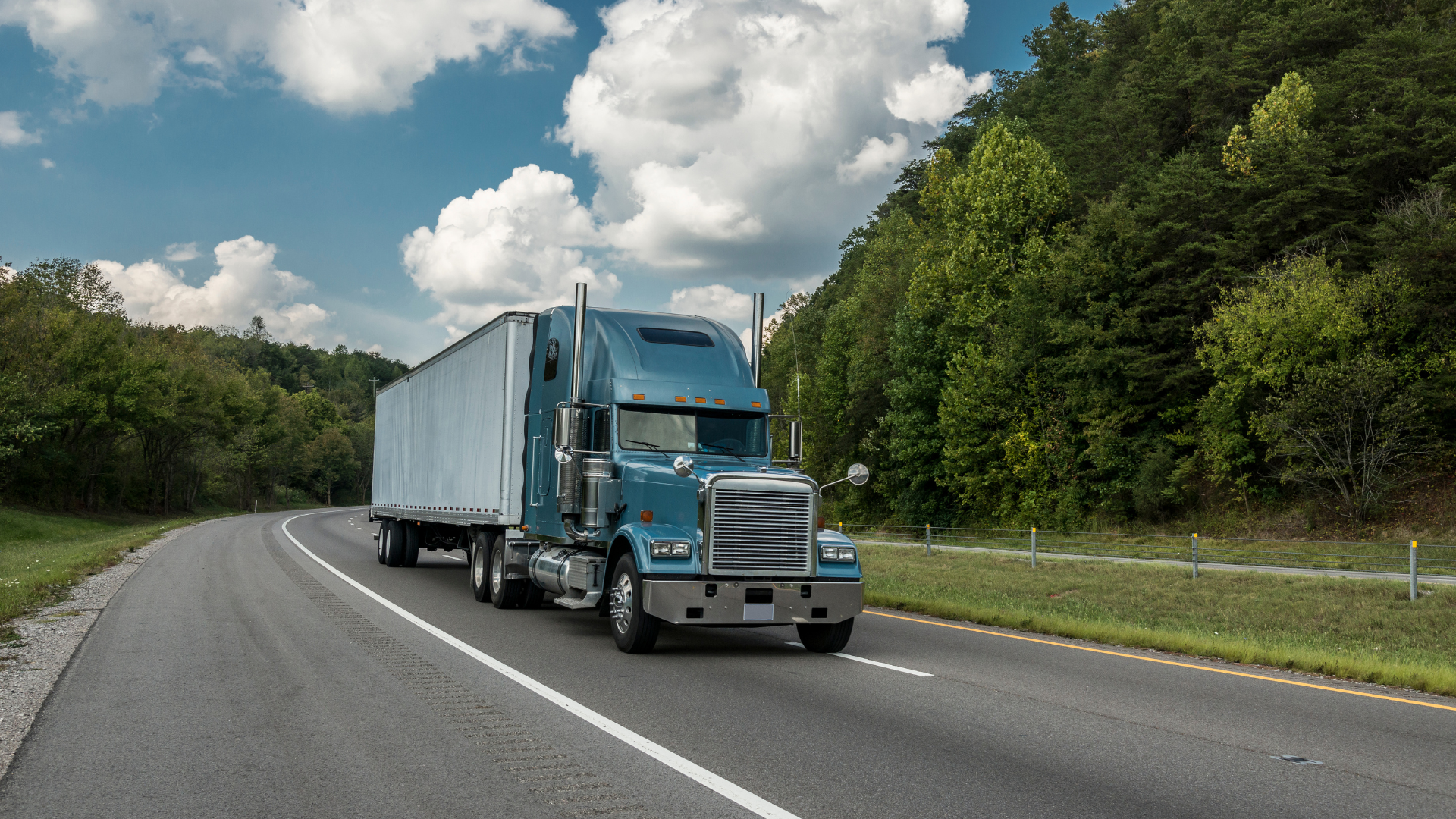 Blue semi-truck driving on a winding road through a lush, green landscape under a cloudy sky.