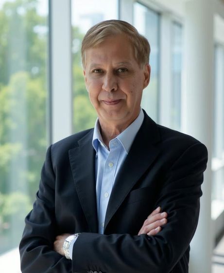 Man in suit with arms crossed, standing near a window.