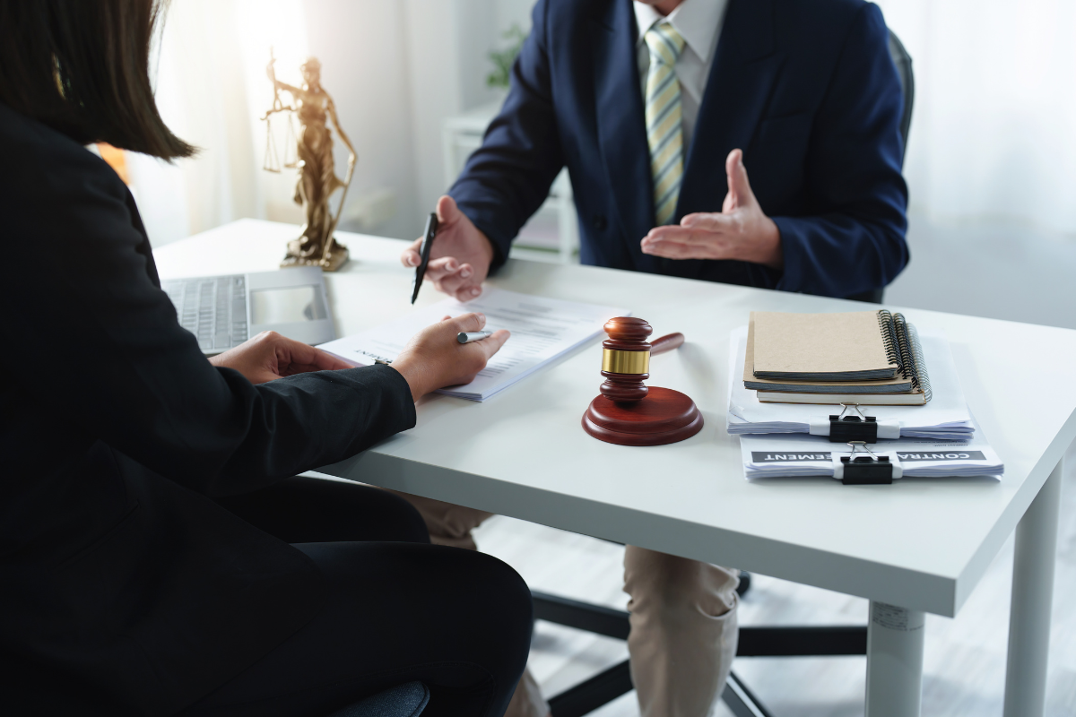 Lawyer explaining document to client at a desk. Gavel, statue of justice, and files present.