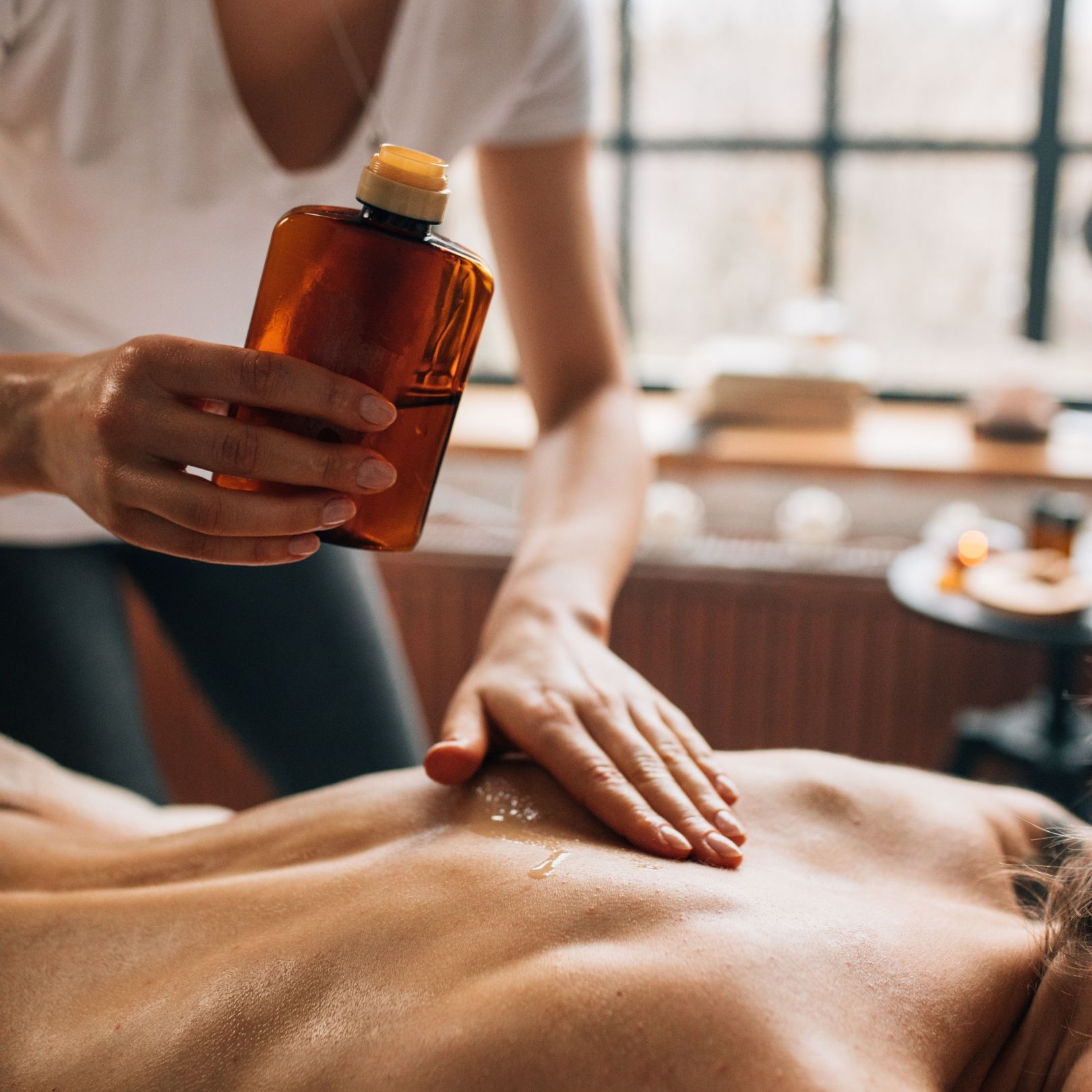 Massage therapist pouring oil onto a person's back; hands smoothing oil, sunlight, warm setting.