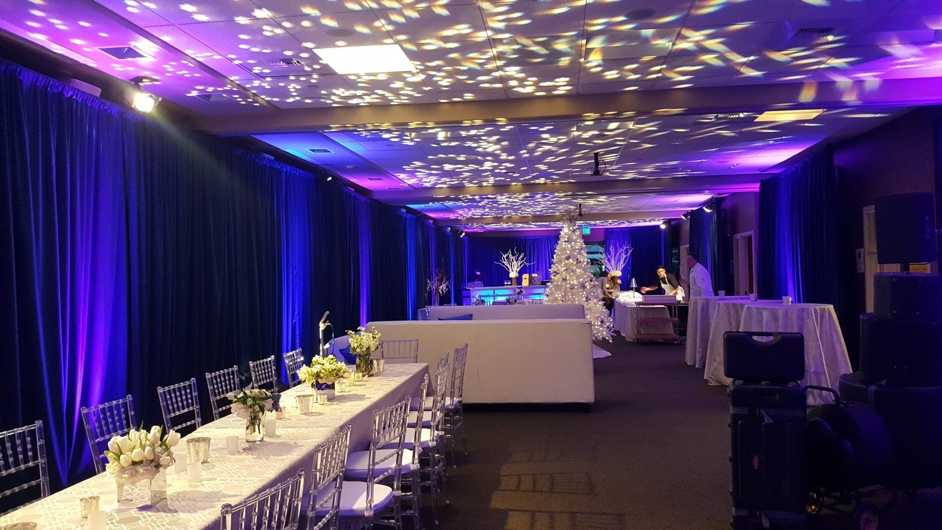 Event space with tables and chairs set for a party, blue and purple lighting, and a white Christmas tree.