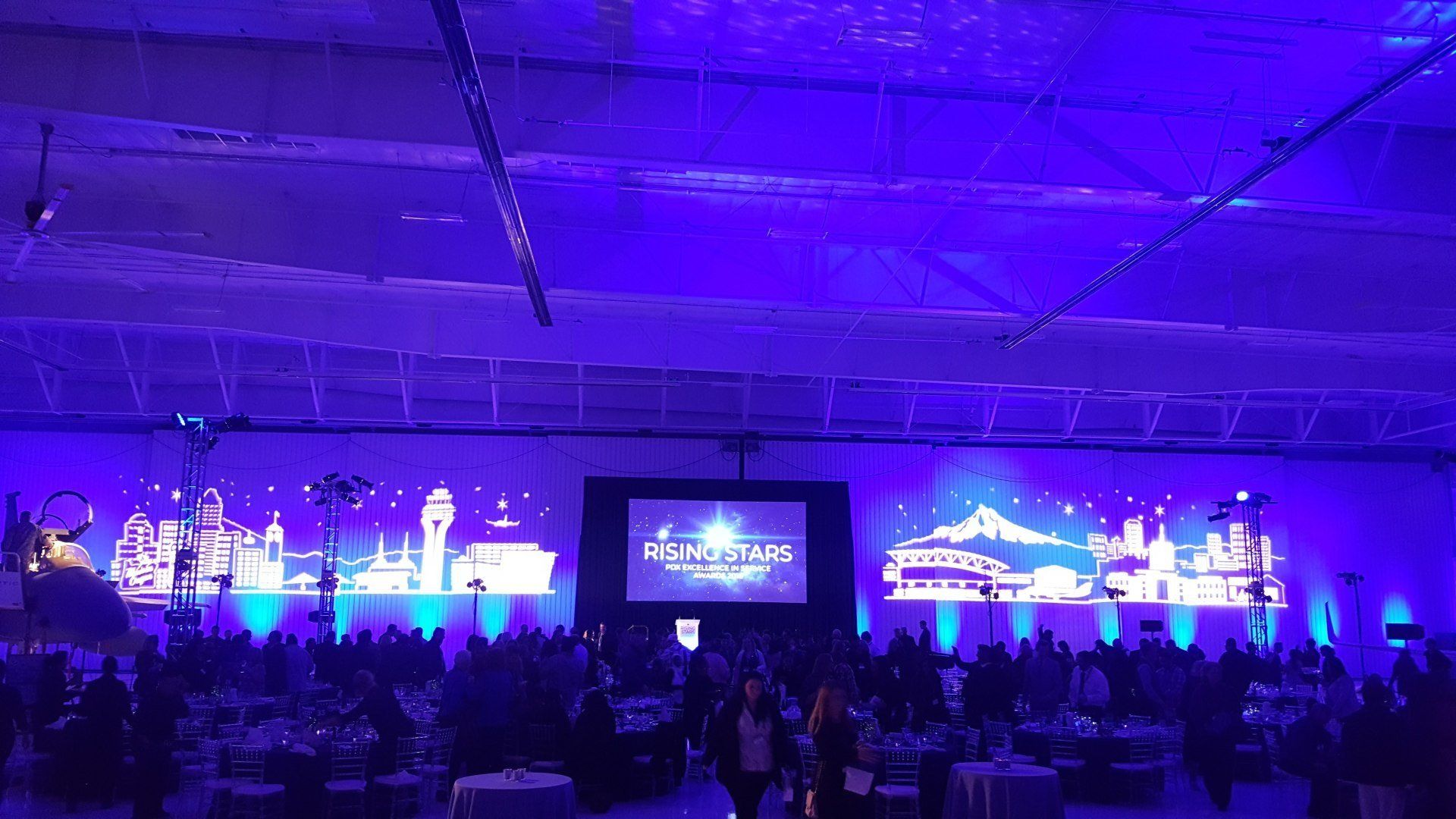 Large event hall with blue lighting, a stage, and seated attendees at tables.