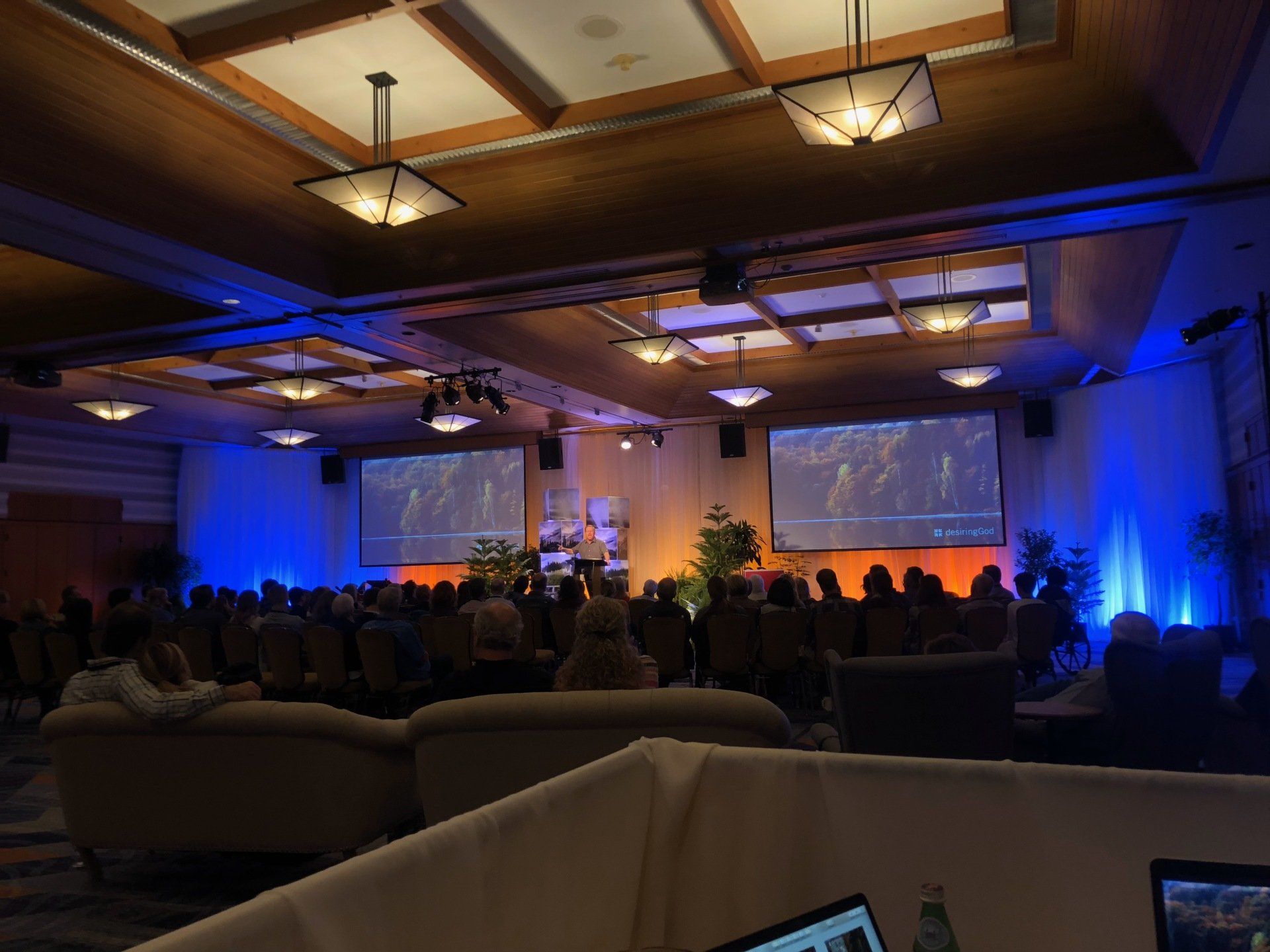 Conference hall with audience facing stage with screens and presenters. Blue and orange lighting.