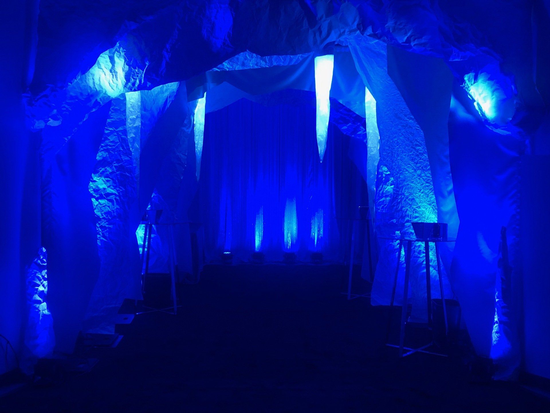 Blue-lit tunnel entrance with ice stalactites, draped white fabric, and a dark pathway ahead.