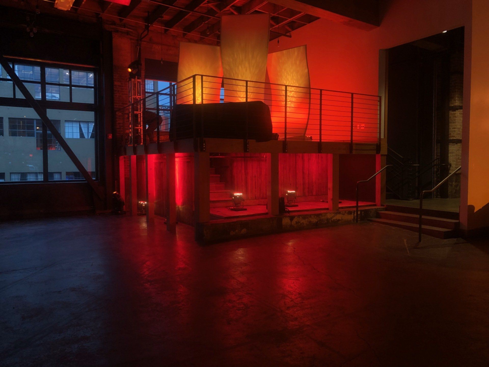 Empty venue with red lighting on a stage with a bar, stairs, and windows.