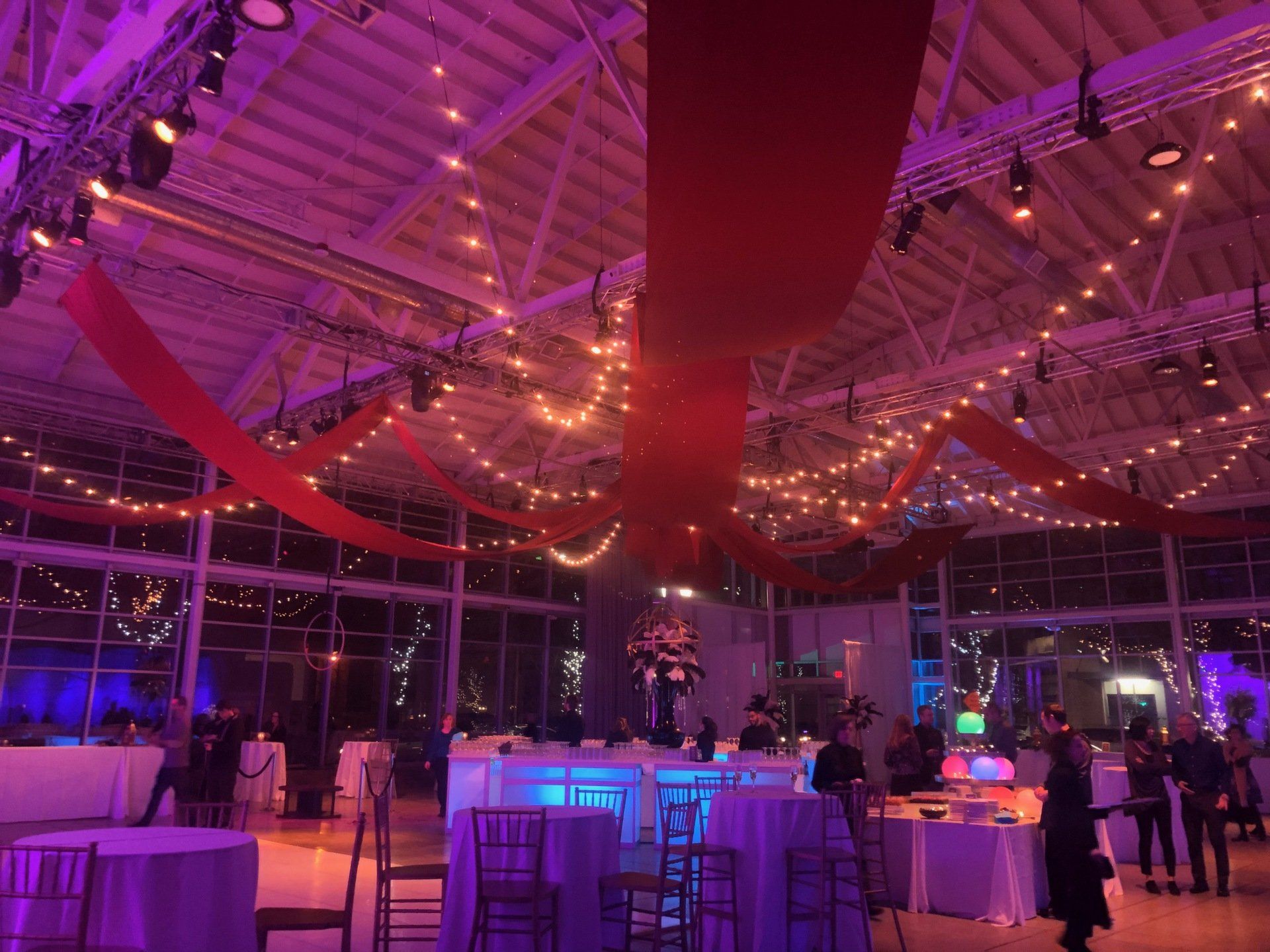 Indoor event space with tables, string lights, red fabric, and purple lighting.
