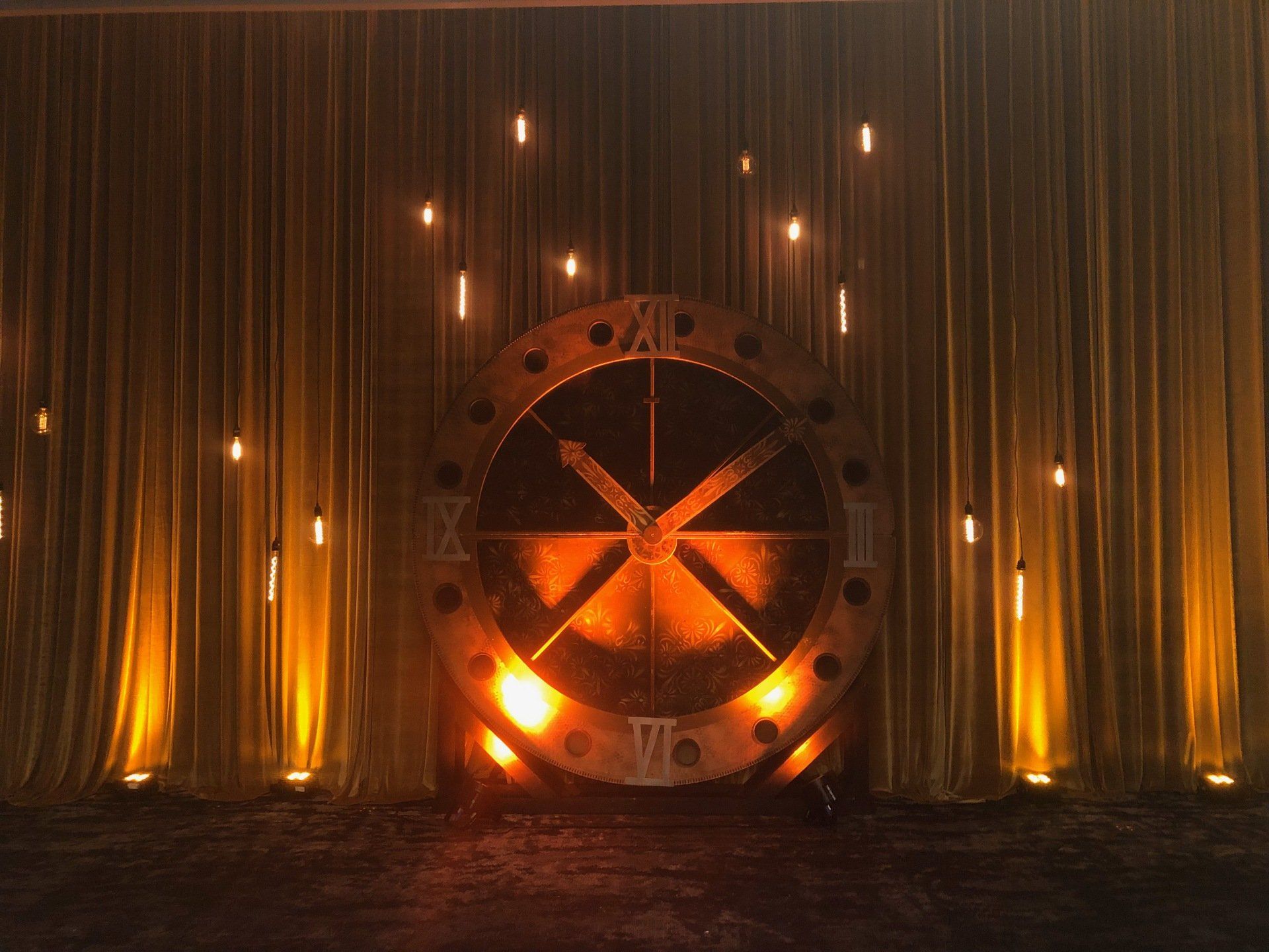 Large clock with roman numerals, glowing lights, golden curtains. Dark, dramatic setting.