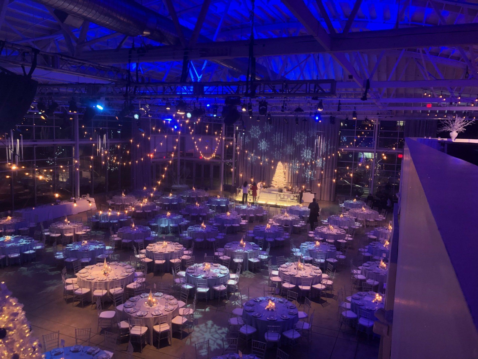 Blue-lit event space with round tables set for a gathering; strings of lights and a stage in the background.