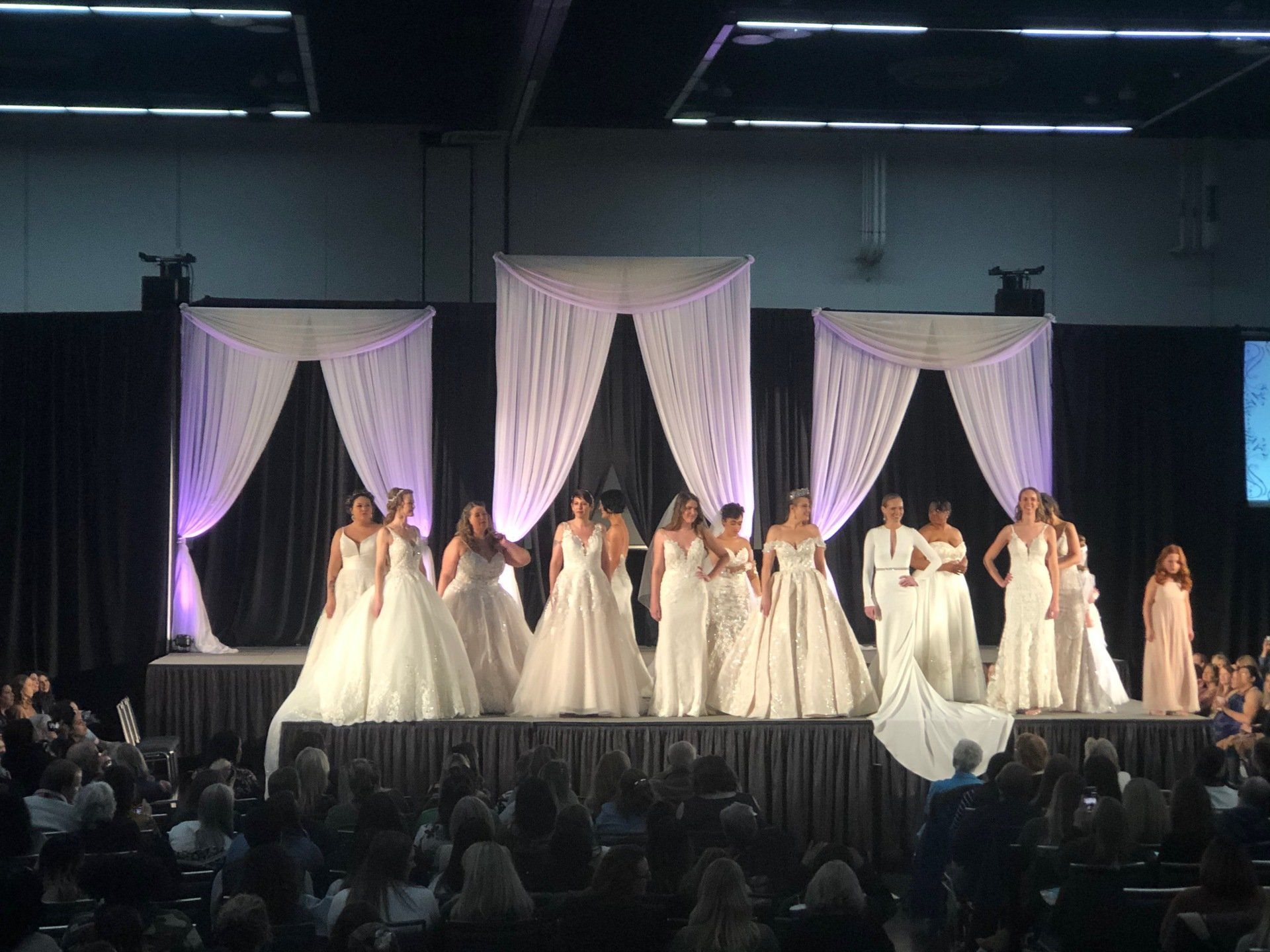 Brides wearing white dresses on a stage with curtains, in front of an audience.