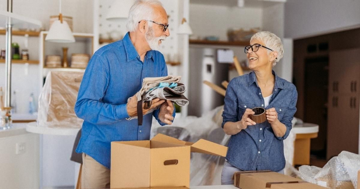 Elderly couple packing boxes in a brightly lit kitchen; man holding clothes, woman holding items.