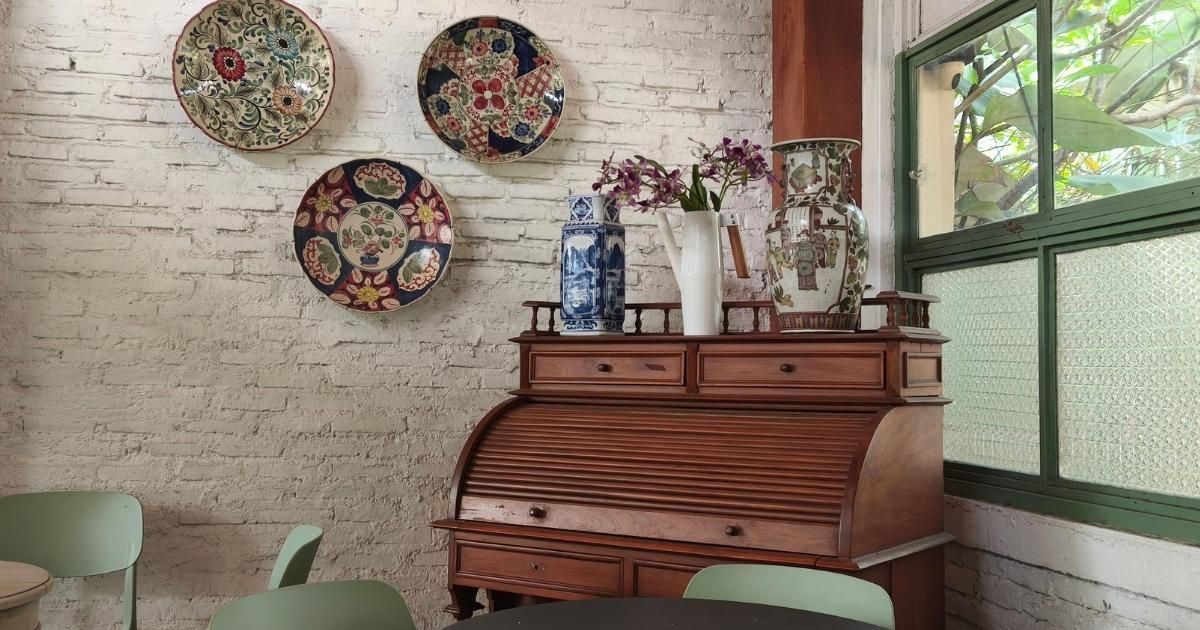 Interior with antique desk, decorative plates on brick wall, vase of flowers, and a window.