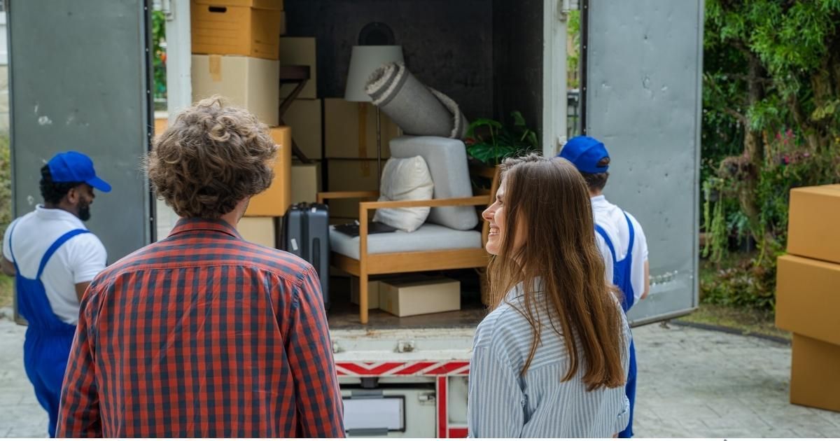 Couple watches movers load a truck with furniture and boxes. Men in blue uniforms. Outdoors.