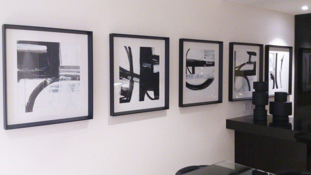 Five framed abstract black and white artworks on a white wall, above a dark cabinet with black decorative objects.