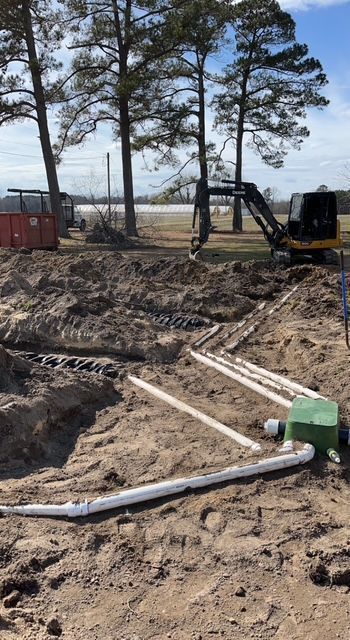 A construction site with a lot of pipes in the dirt and trees in the background.