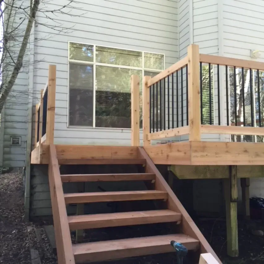Wooden deck with stairs leading down, built against a house with light blue siding. Black metal railings.