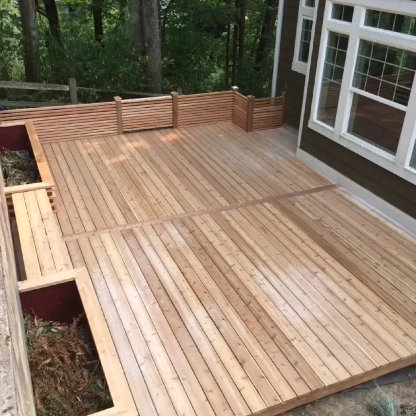 Wooden deck with slatted fence, beside a brown house and green trees.