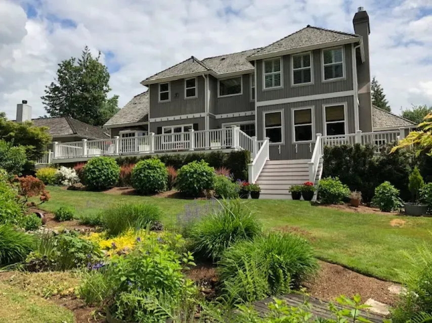 Gray two-story house with white trim and multi-level decks, surrounded by lush green garden with flowers.