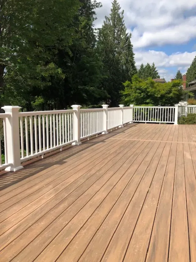 Wooden deck with white railing, surrounded by trees under a blue sky with clouds.