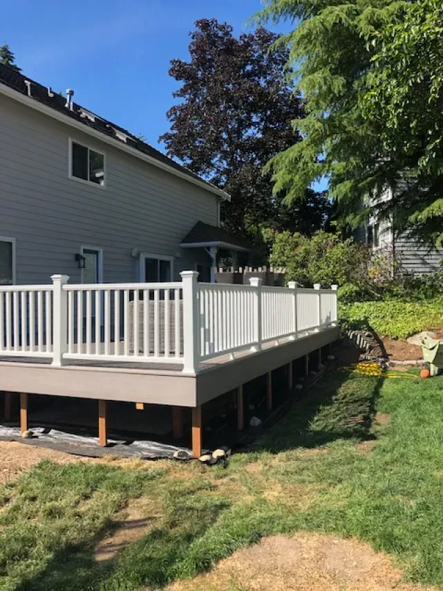 White railing surrounds a gray-decked deck attached to a gray house, set in a yard.