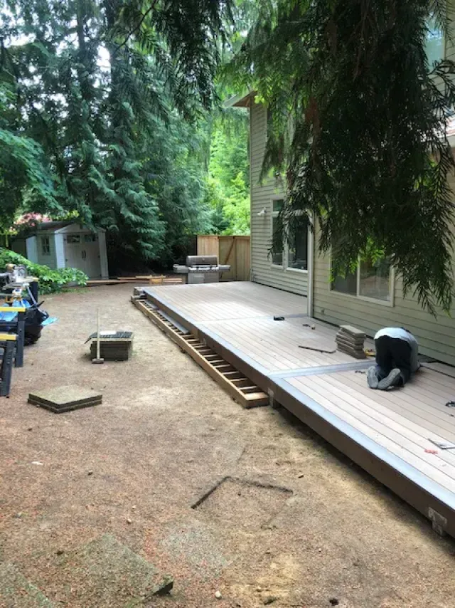 Backyard with partially constructed deck, shed, and someone working. Brown and green tones dominate the scene.