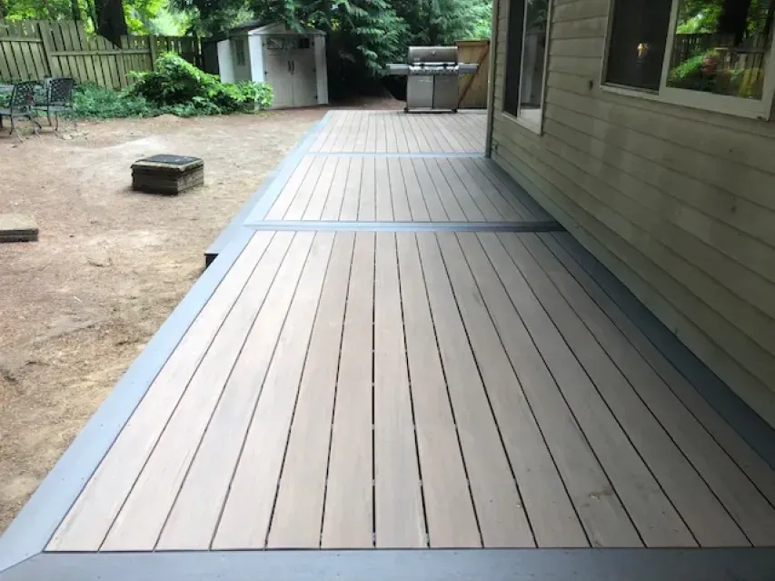 Wooden deck with gray border extending from the side of a house into a backyard.