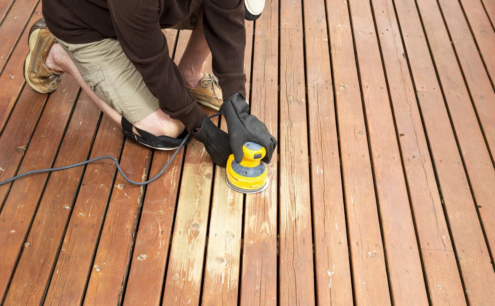 Person sanding a wooden deck with a power sander; brown deck boards and khaki pants visible.