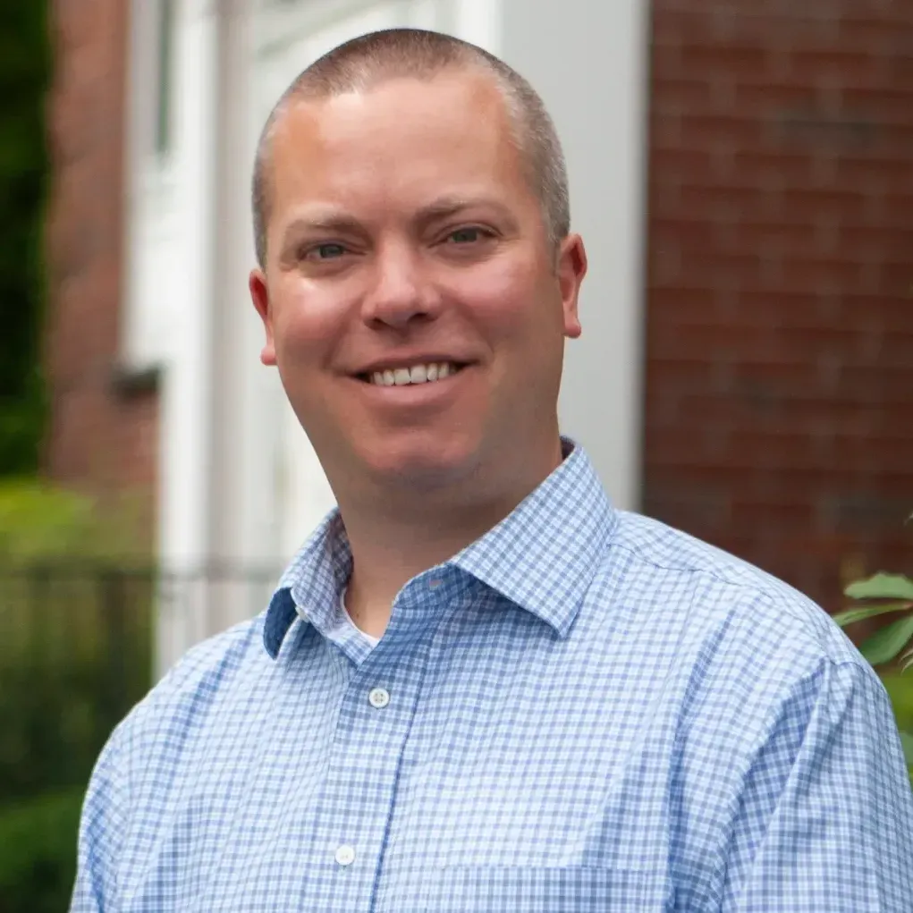 A man in a blue plaid shirt is smiling in front of a brick building.