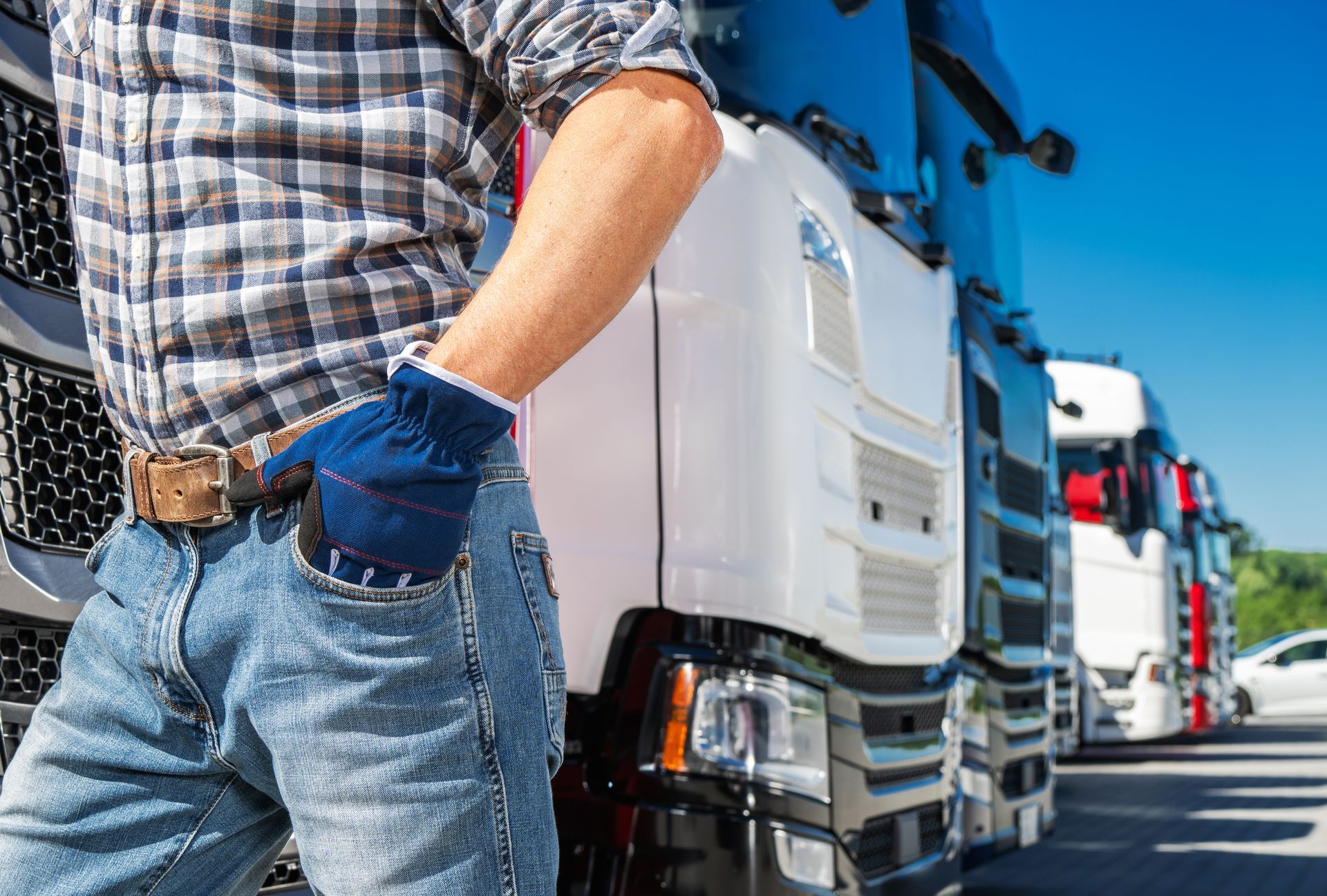 Truck driver with hand in pocket, standing in front of a line of semi-trucks on a sunny day.