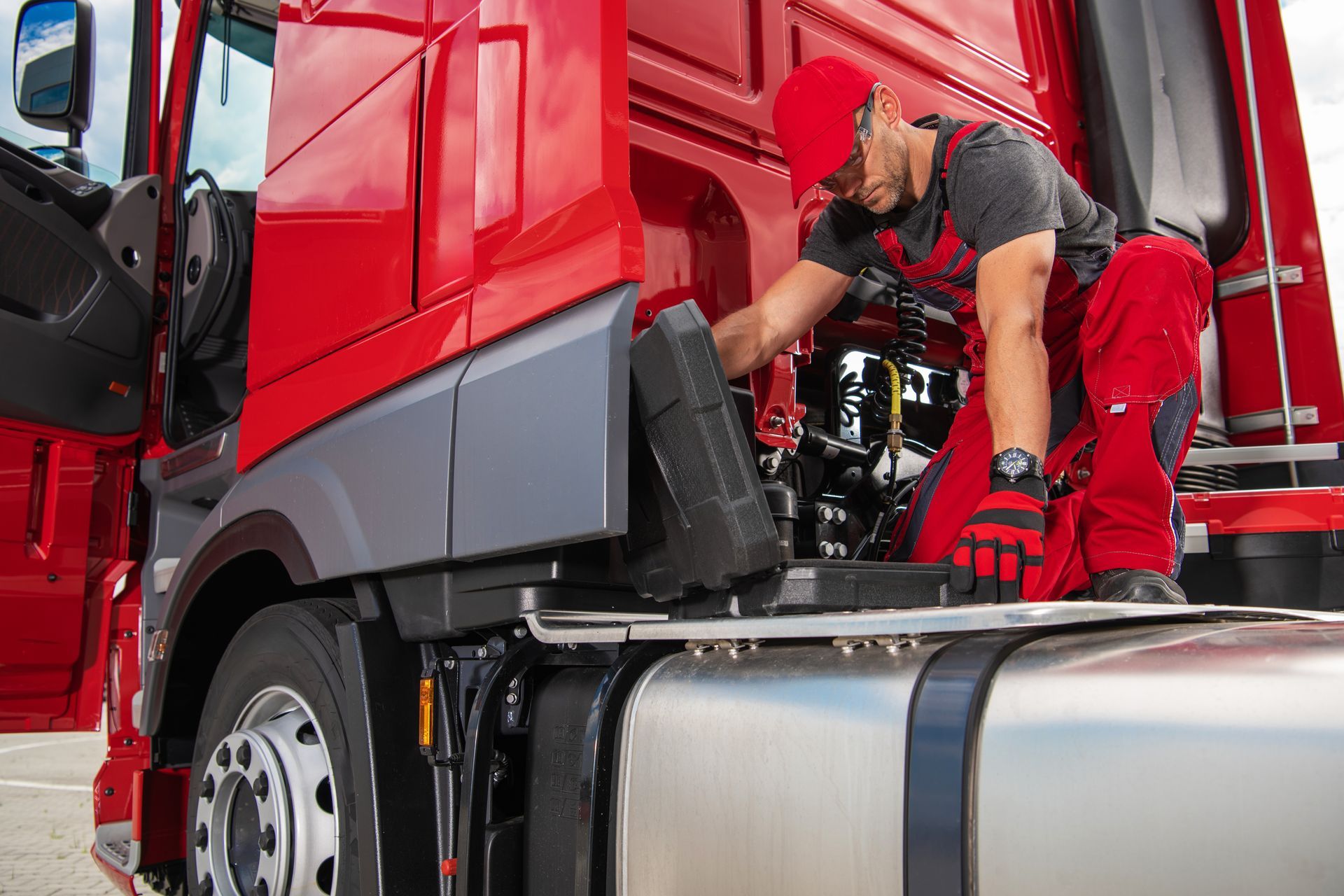 Mechanic in red overalls and hat working on the engine of a red semi-truck.