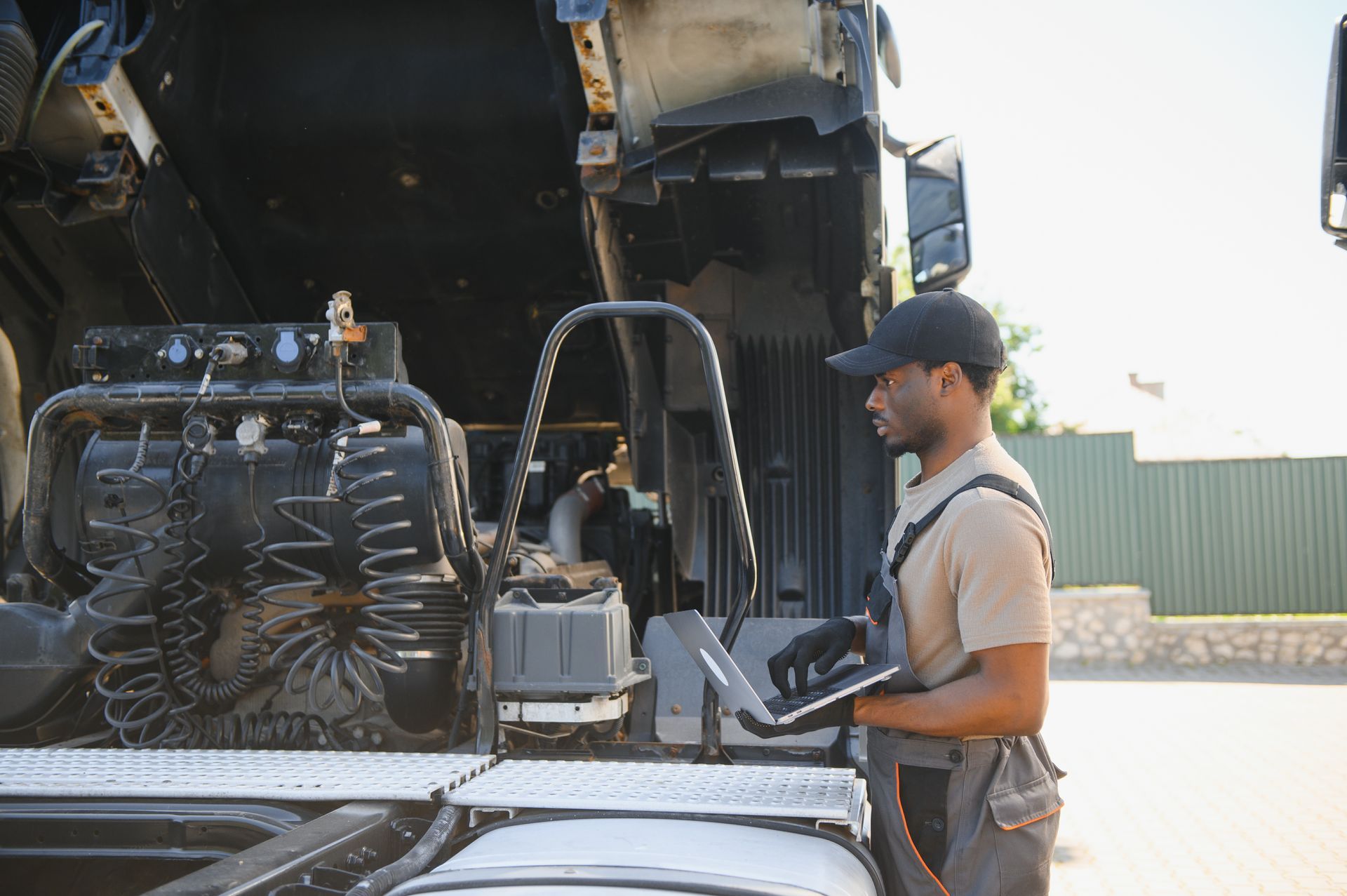 Mechanic uses a laptop to diagnose a semi-truck engine. Outdoors, sunny.