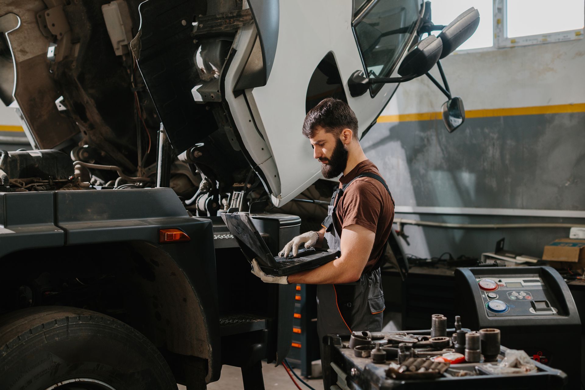Mechanic in a garage, using a laptop while inspecting a vehicle with an open hood.