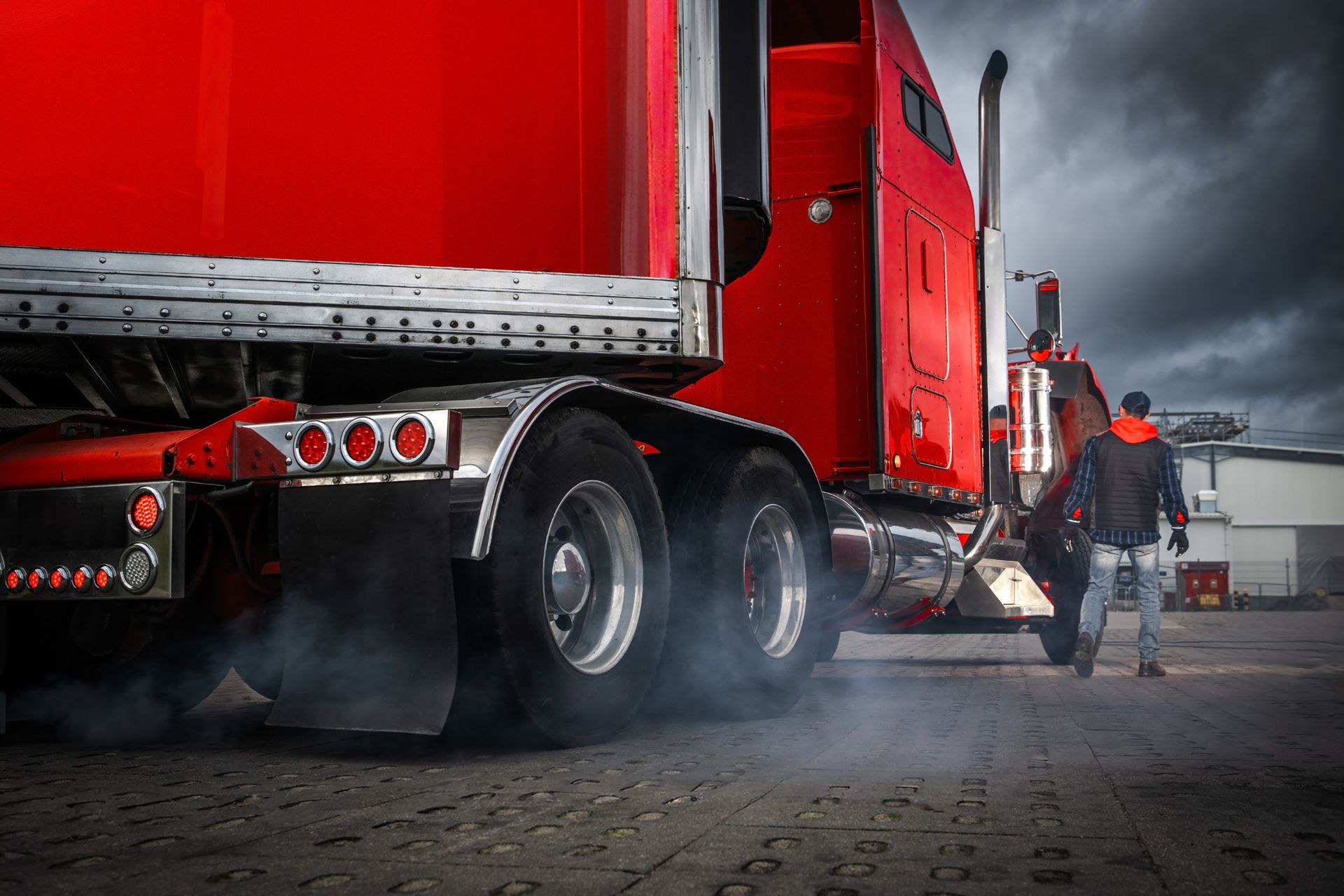 Red semi-truck with exhaust, person walking towards it on paved surface under a cloudy sky.