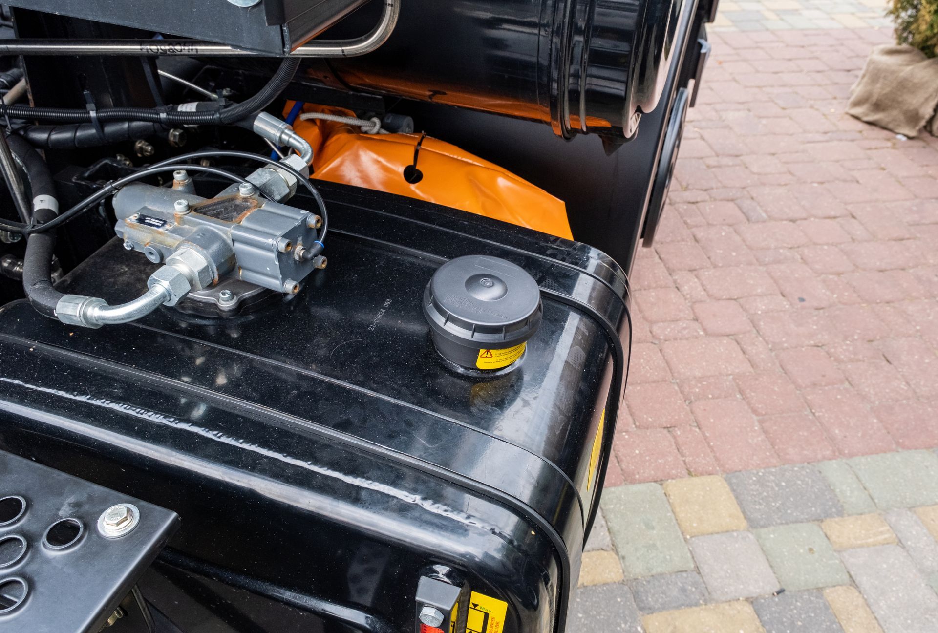 Black machinery with a fuel cap on top, hydraulic components visible, and an orange container in the background.