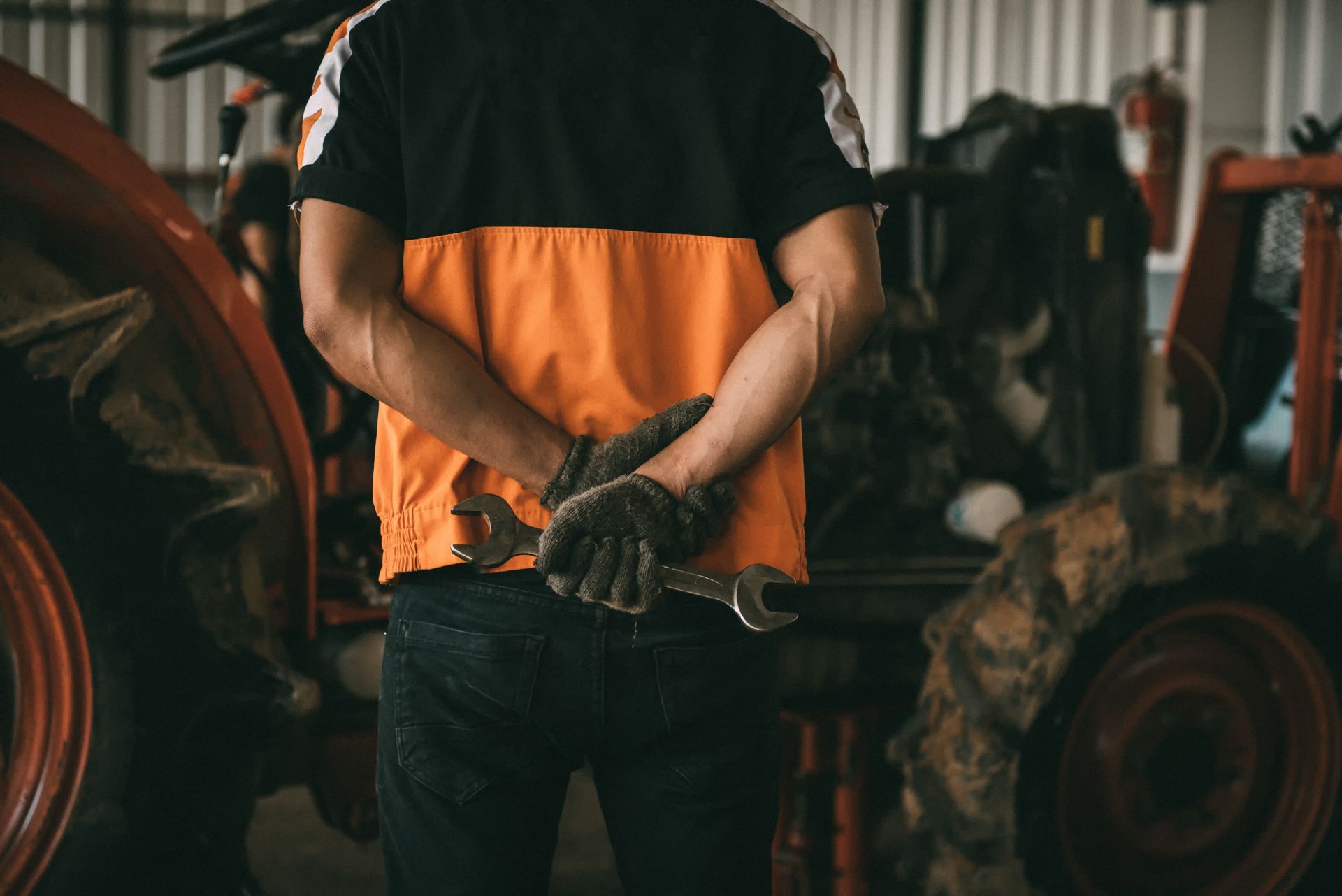 Mechanic holding a wrench, hands clasped behind back. Wearing black and orange work shirt, in a garage.