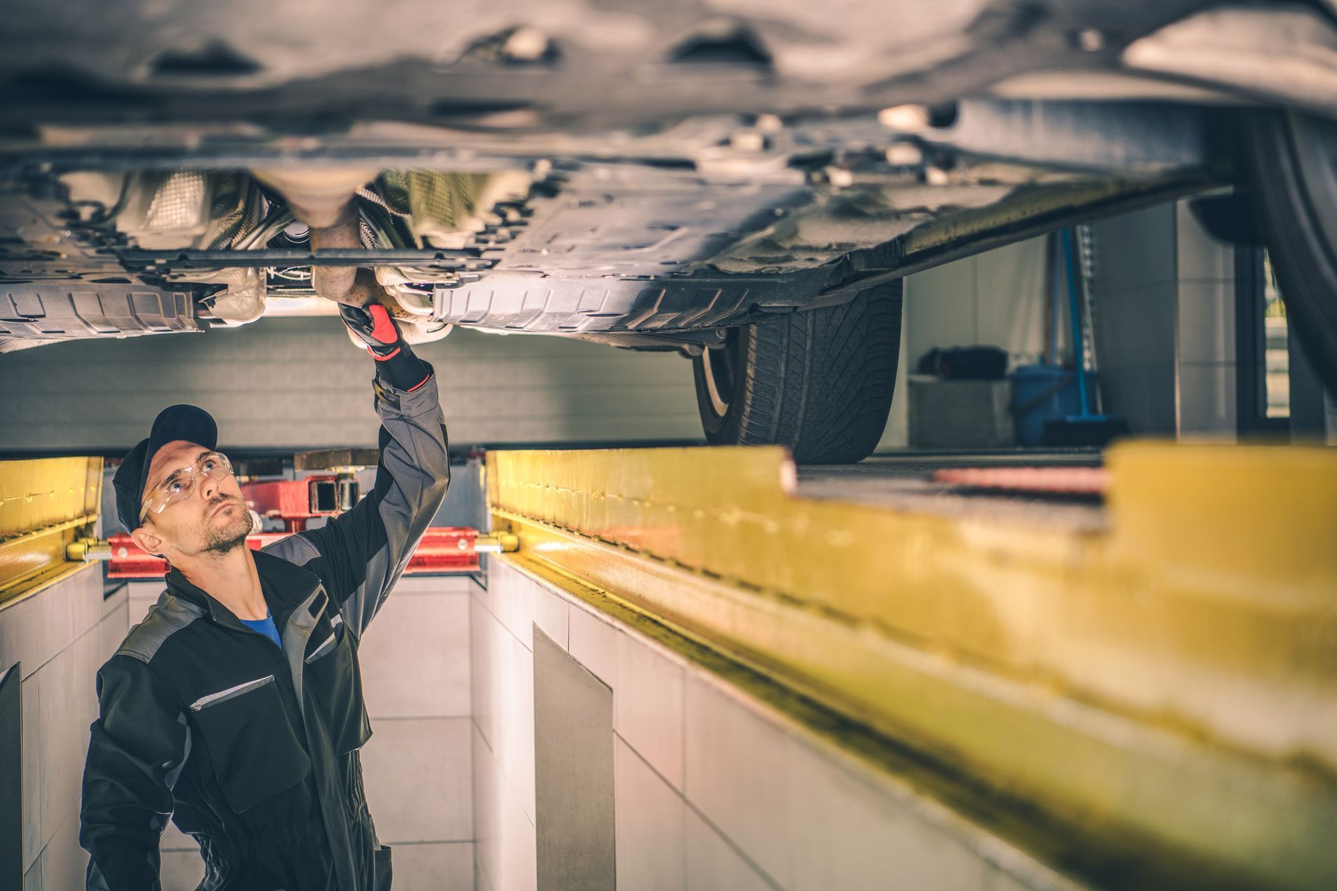 Mechanic in dark work clothes inspects a car's undercarriage in a garage, using a flashlight.