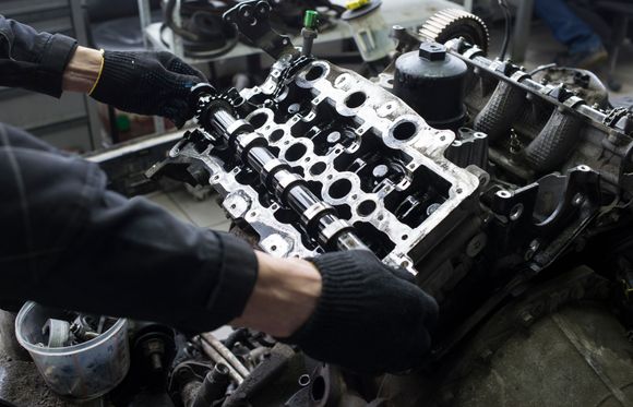 Hands wearing black gloves working on an engine cylinder head in a garage.