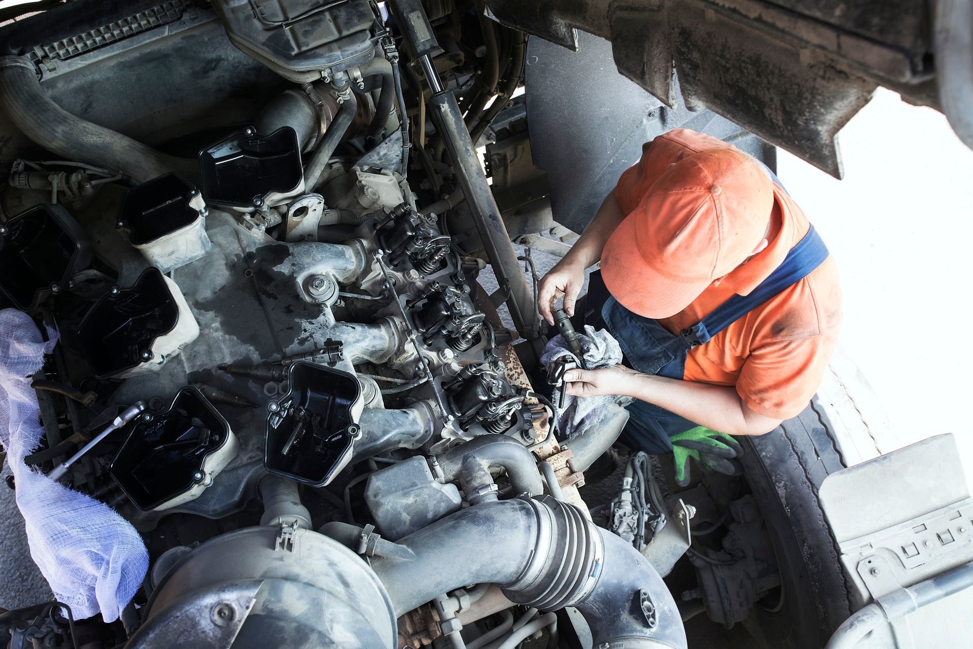 Mechanic in orange shirt and overalls working on a truck engine, viewed from above.