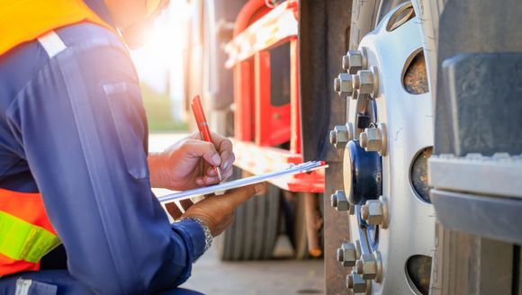 Person in safety vest inspecting a truck wheel, writing on a clipboard.