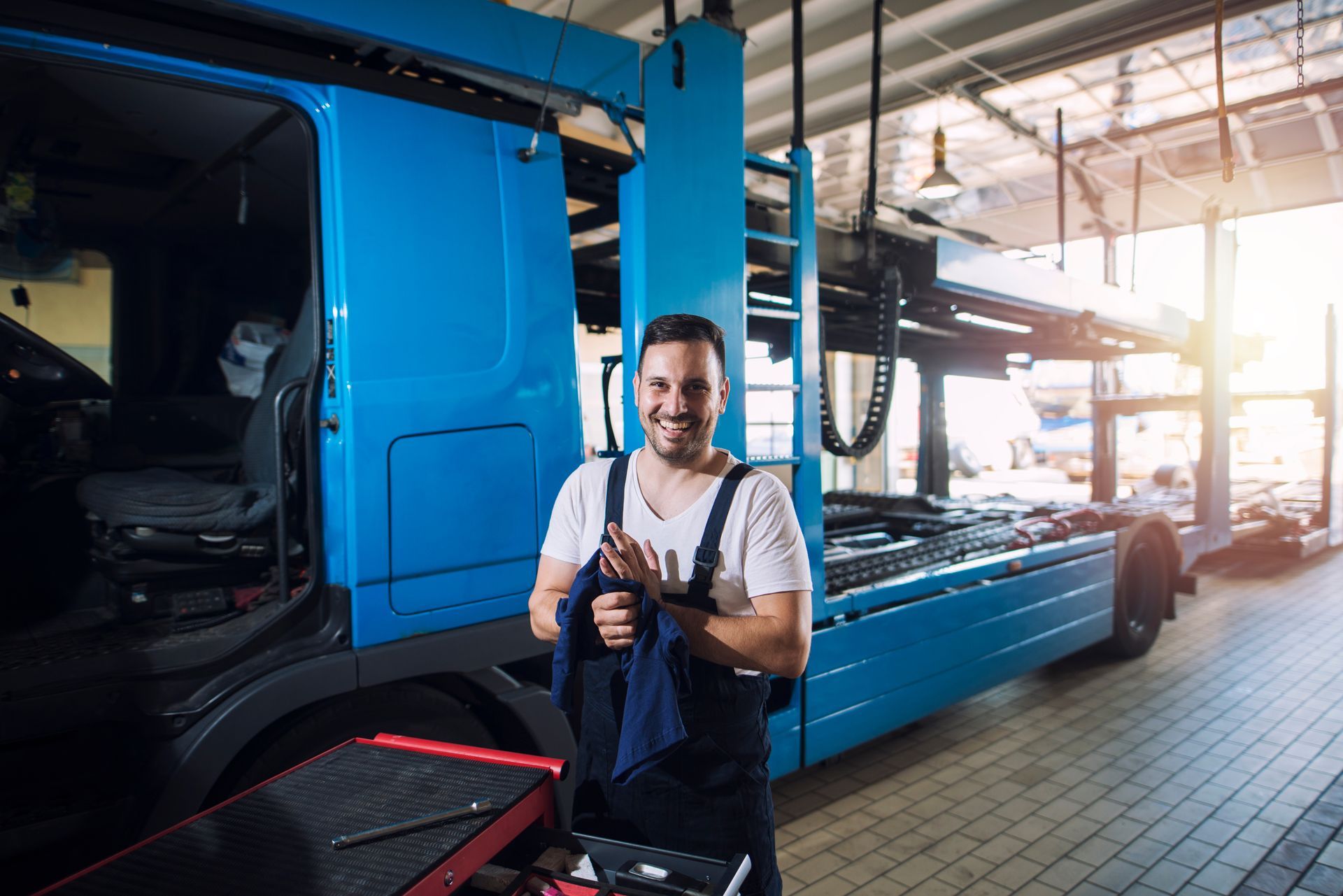 Mechanic stands smiling, holding a rag, beside a blue truck in a repair shop.