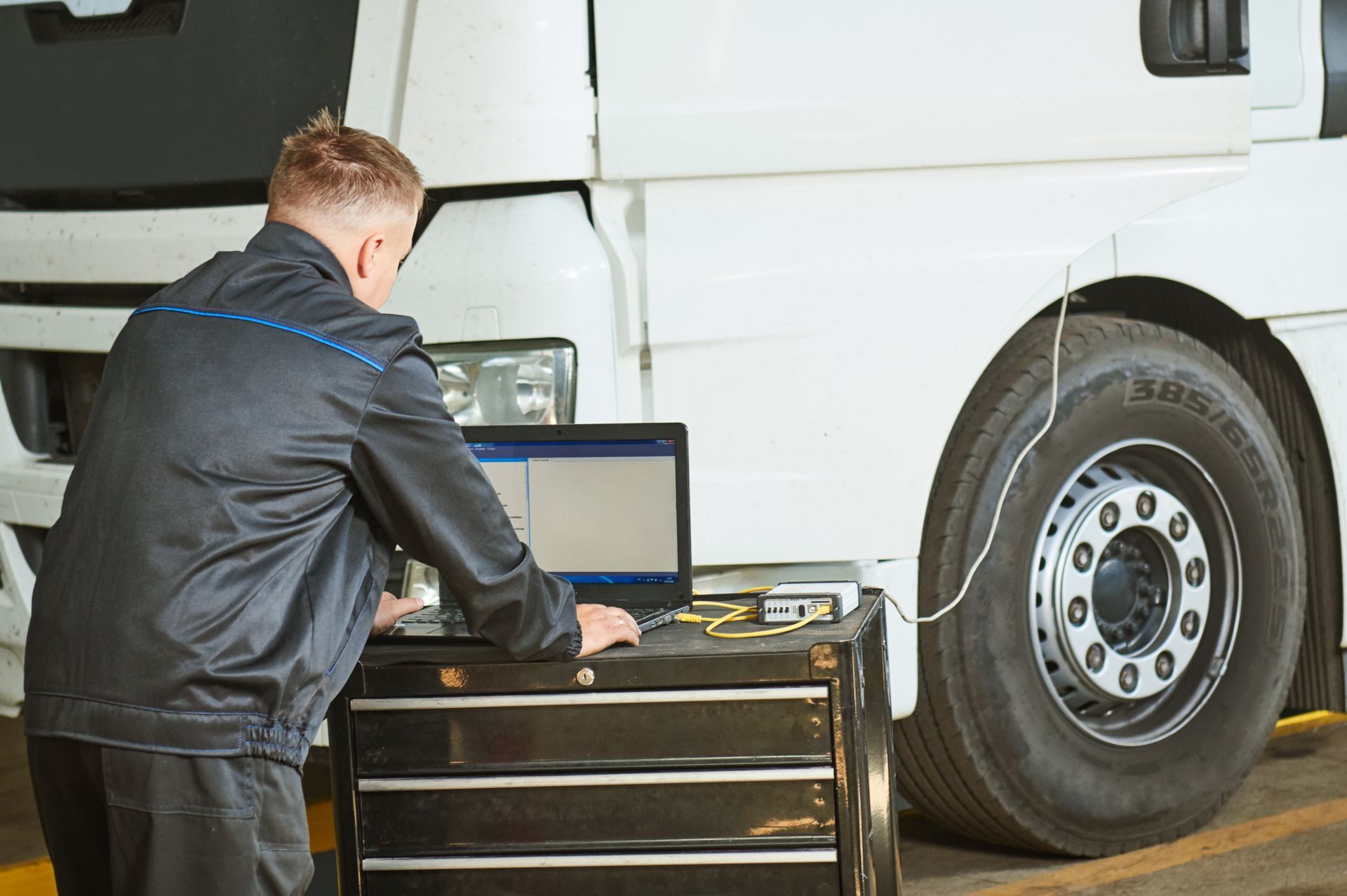 Mechanic using laptop to diagnose a truck tire, parked in a garage.