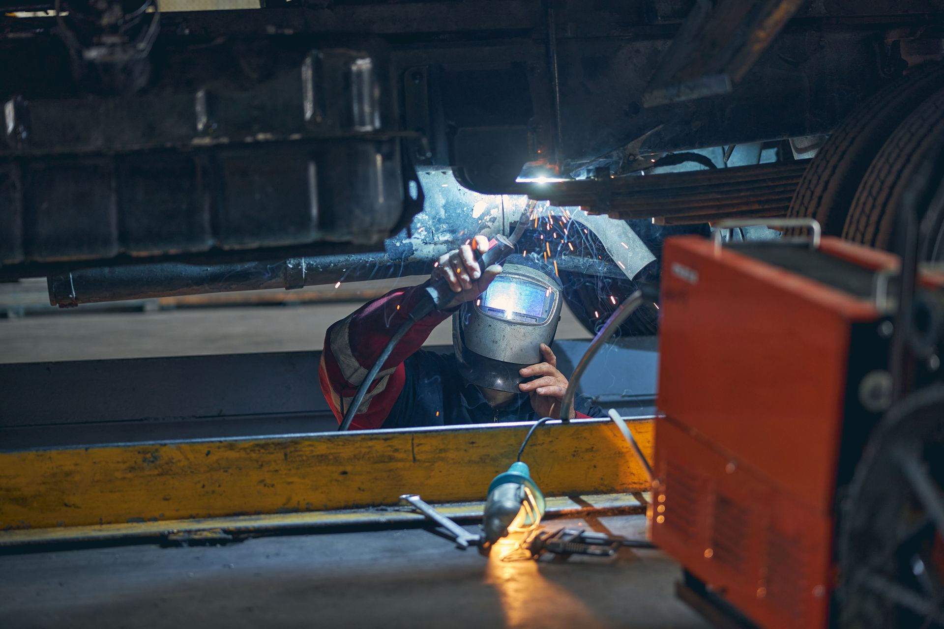 Welder in a workshop using a torch, sparks flying, with welding equipment visible.