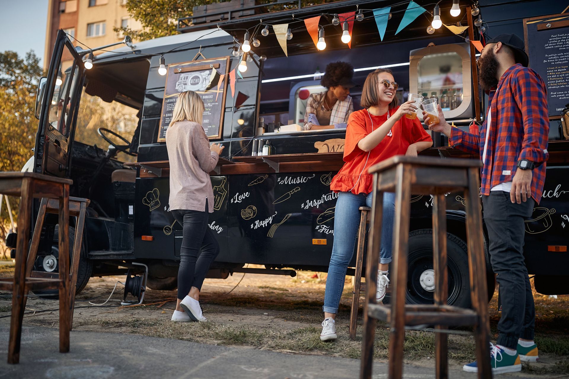 People at a food truck, clinking glasses. Outdoor setting with string lights and tables.