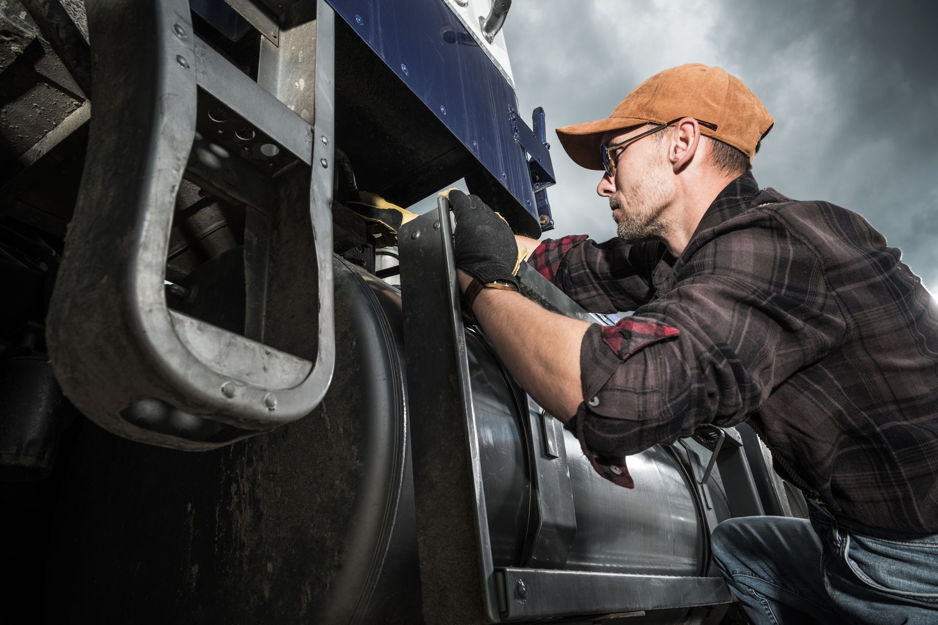 Man in cap and gloves working on a large truck's exterior; outdoor setting.