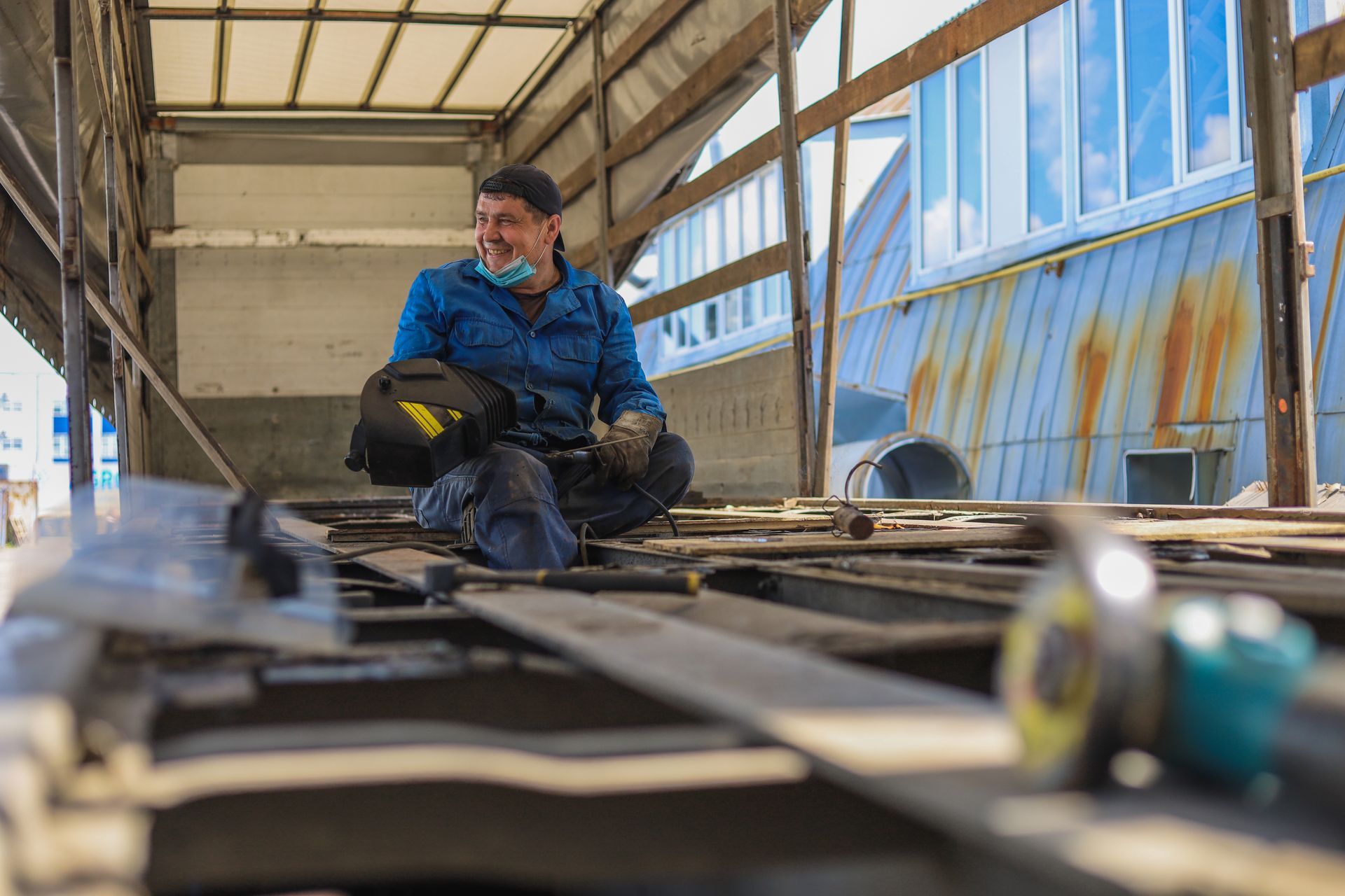 Man in blue overalls smiles, holding welding helmet, sits inside metal structure.