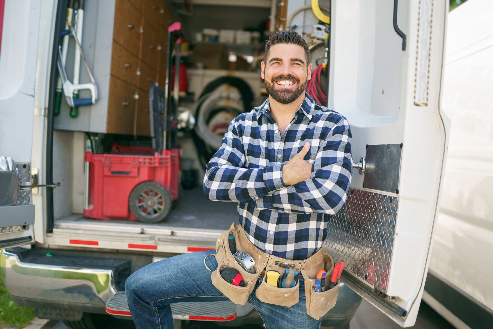 Man with tool belt smiles in front of a van with open door, tools visible inside.