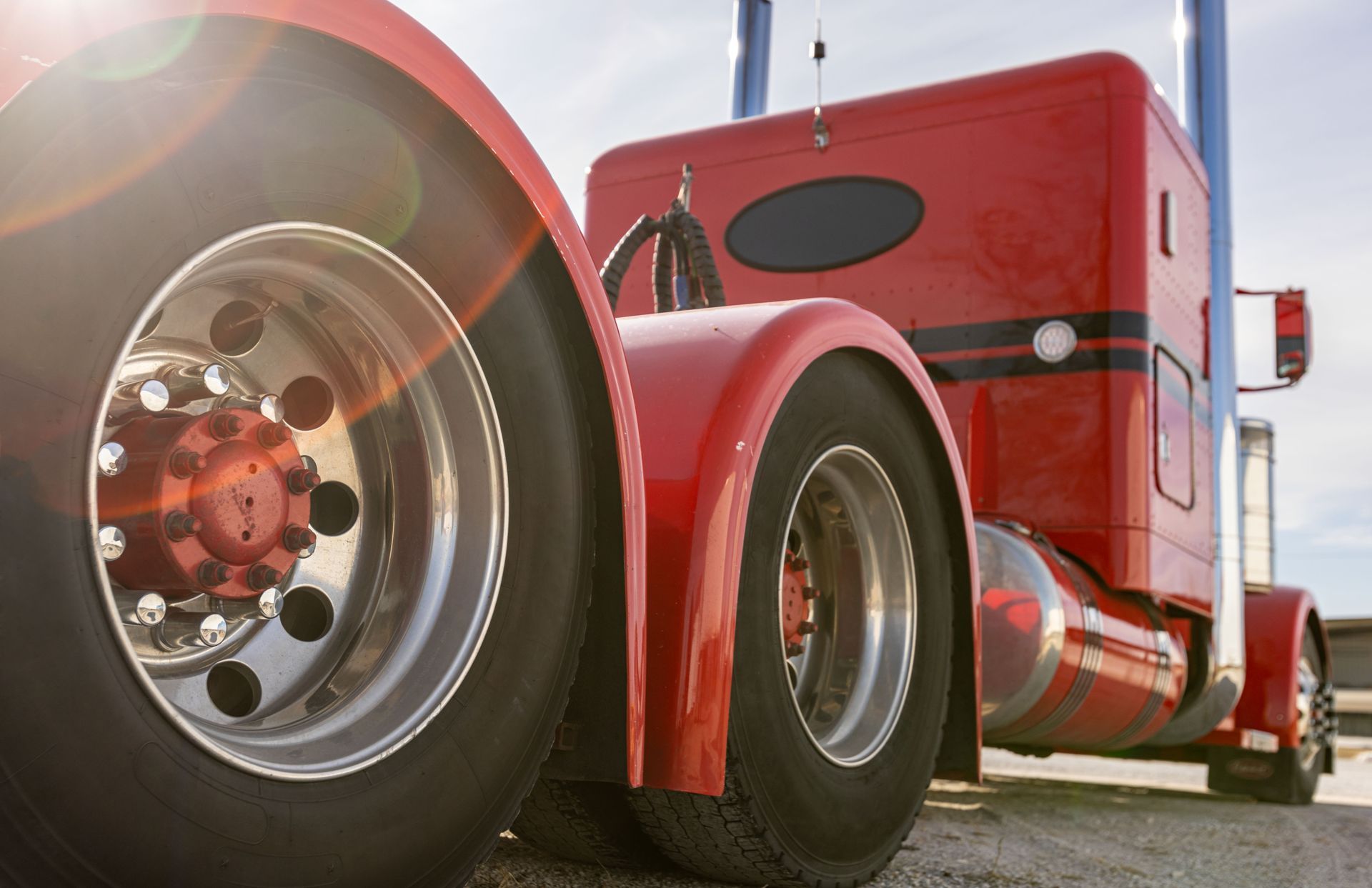 Red semi-truck rear tires and cab, chrome wheel details, bright sunlight.